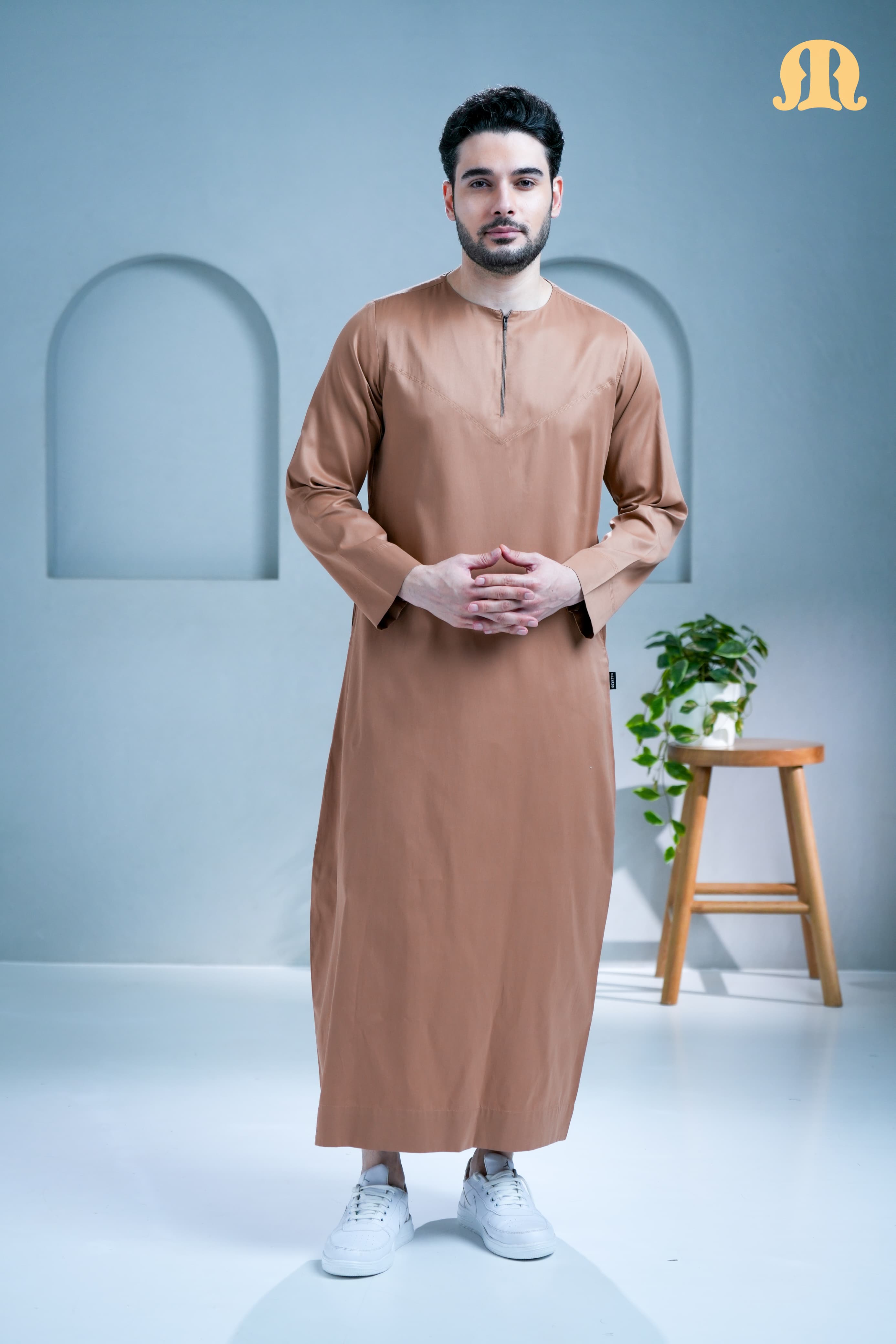 Man wearing a brown traditional outfit standing in a minimalistic room with a plant and stool.
